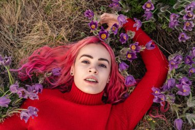 A young woman with vibrant pink hair lies peacefully in a meadow surrounded by purple flowers. Her bright red sweater contrasts beautifully with the natural backdrop.