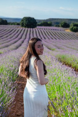 A woman in a white dress strolls through a sprawling lavender field, smiling under a bright, clear sky. The lush purple blooms stretch into the distance