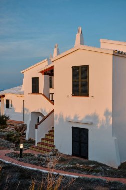 Charming traditional houses in Arenal des Castell, Menorca, bask in the warm glow of the sunset, showcasing the region's unique architectural style and serene coastal atmosphere.