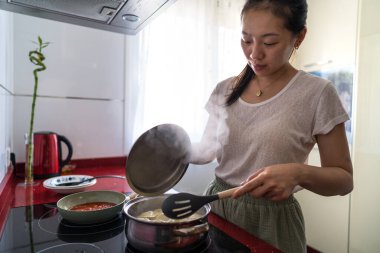 A woman in a modern kitchen prepares pasta with tomato sauce, embodying a casual home lifestyle. The scene captures real-life cooking in an everyday setting