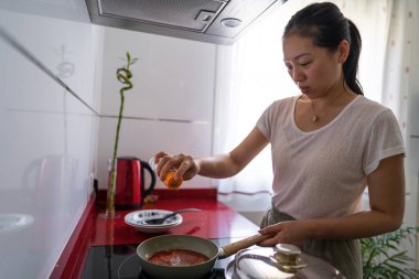 Woman cooking at home in a modern kitchen, using spices to flavor her tomato sauce. Casual home lifestyle captured perfectly, blending culinary creativity with everyday comfort