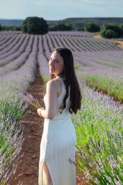 A woman walks joyfully through a vibrant lavender field on a sunny day, holding a small bouquet. The scenery evokes a sense of tranquility and natural beauty