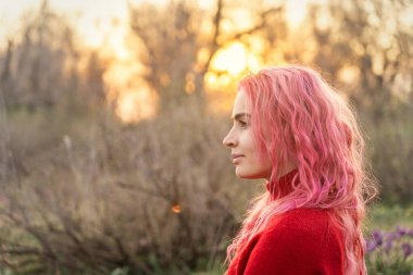 A young woman with vibrant pink hair stands peacefully in a natural setting, surrounded by flowers and bathed in warm, golden sunlight, embodying serenity and beauty.