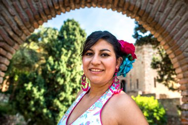 Woman dressed in vibrant flamenco attire stands gracefully in a historic Andalusian garden with a Moorish castle backdrop, celebrating cultural authenticity and tradition