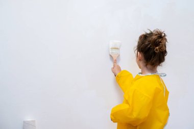 Back view of unrecognizable little girl in a yellow protective coat painting a room with a brush. She adds fresh, creative touches to the walls, showcasing youthful enthusiasm.