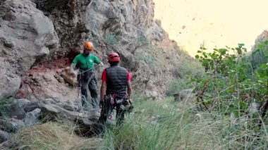 Two men in helmets prepare ropes for traditional climbing in a rocky canyon. One climber organizes gear with hexes and camalots, while the other observes. The setting is lush and rugged.