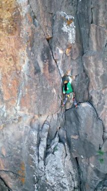 Back view of unrecognizable man climb a rugged cliff, equipped with climbing gear, camalots, and protective helmet. The natural rock face is the main background.