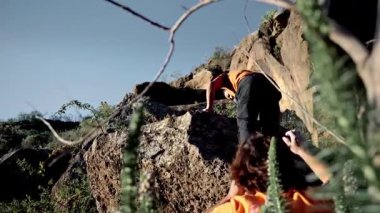 Low angle back view of two men prepare for a traditional climbing session, with one warming up on easy rocks while the other spots for safety. They are surrounded by nature and gear up for a bigger climb.
