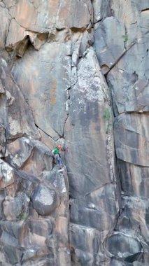 A climber clad in a helmet and harness scales a rugged cliff using traditional climbing gear. The scene captures the precision of placing camalots and anchors on a rocky surface.