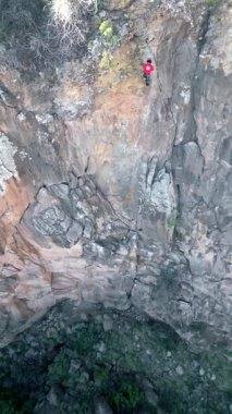 From above two men engage in traditional climbing on a steep rocky cliff, showcasing teamwork and skill in an outdoor setting. They use ropes and climbing gear for safety.