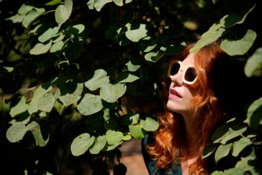 Fashionable redhead woman wearing sunglasses and enjoying nature under green leaves in a sunny day