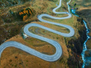 Sonbaharda günbatımında Alp Vadisi 'ndeki nehir, dolambaçlı dağ yolunun hava manzarası. İsviçre dağ geçidindeki yol manzarası, sarı çimenler, sonbaharda ağaçlar. Renkli manzara. İsviçre 'de Alpler