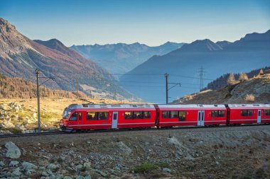 İsviçre 'nin İsviçre Alpleri' ndeki yüksek dağlık bir vadide gün batımında kayalık yamaçlar, çam ağaçları ve katmanlı dağ siluetleriyle seyahat eden Kızıl Bernina Ekspresi treni. Sonbahar