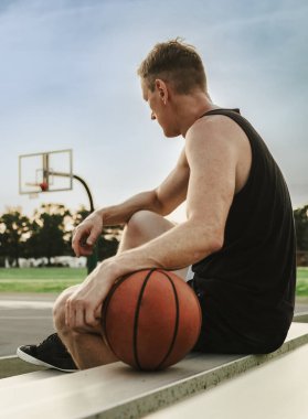 A man sitting and leaning on a basketball while looking at the court in the distance during the evening in the United States.