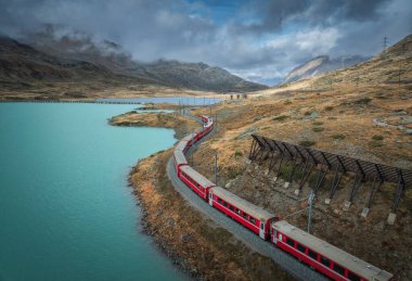 İsviçre Alpleri 'ndeki turkuaz göl boyunca kıvrılan kırmızı Bernina Ekspres treni, kayalık tepeler ve sonbahar dağları manzarasında dramatik bulutlu gökyüzü. Sonbaharda trenin üst görüntüsü