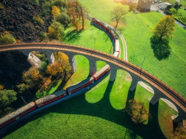Güneşli bir sonbahar gününde İsviçre 'de, Brüksel Viyana' dan geçen kırmızı Bernina Ekspres treninin hava aracı görüntüsü, renkli ağaçlar, dağ vadisi manzaralı dağlık bir köy. Alplerdeki trenin üst görünümü