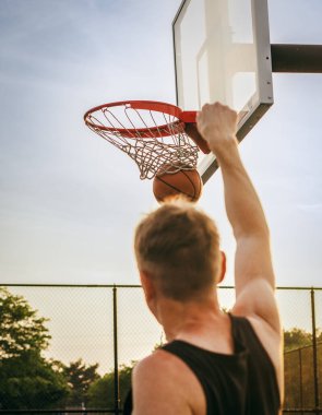 A man seen from behind throws a basketball that lands in the hoop against the sky in the United States.