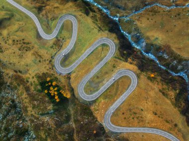 Sonbaharda günbatımında Alp Vadisi 'ndeki nehir, dolambaçlı dağ yolunun hava manzarası. İsviçre dağ geçidindeki yol manzarası, sarı çimenler, sonbaharda ağaçlar. Renkli manzara. İsviçre 'de Alpler