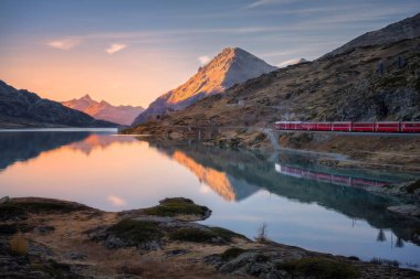 Gün batımında İsviçre Alpleri 'ndeki Bianco Gölü yakınlarındaki Bernina Ekspres treninin parlayan dağ zirveleri, yansımalı su ve kayalık kıyıları. Huzurlu sonbahar manzarası. Sonbaharda dağ treni