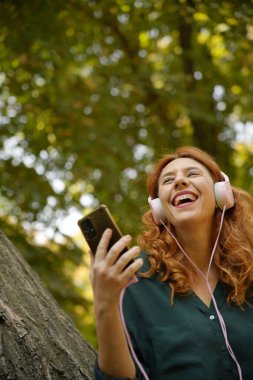 Red-haired woman enjoying music with headphones and smartphone in a forest, expressing joy and relaxation