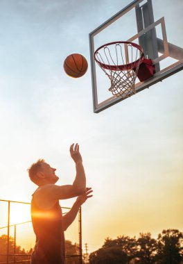 A man throwing a basketball into the hoop from a low angle against a sunset sky in the United States.
