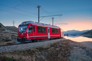 İsviçre 'nin Bianco Gölü yakınlarında gün batımında Bernina Express treni İsviçre Alpleri' nde sakin bir akşam ışığında net yansımalar, dağ siluetleri ve dağlık arazi. Alacakaranlıkta kırmızı tren. Aktarım