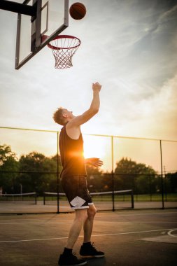 A man throwing the ball from beneath the hoop on an outdoor basketball court during sunset in the United States.