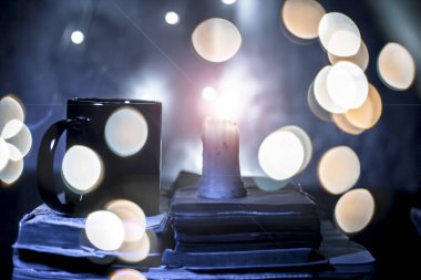 Üzerinde bazı mavi ışık ve bazı kahverengi renkli eski kitap ve bookeh ve Noel ışık üzerinde siyah renkli boş kahve bardağı ile beyaz renkli mum yakın.