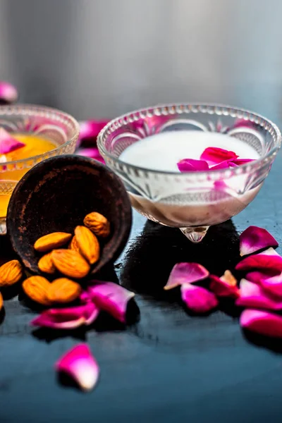 Rose and milk face pack on black colored shiny surface consisting of rose water,almonds and honey in a glass bowl and entire raw ingredients on the surface.Used for instant glow.