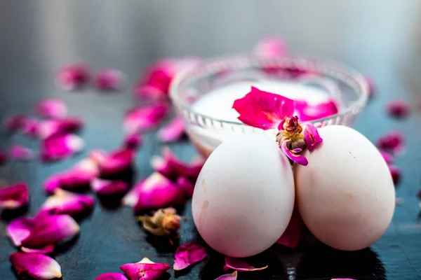 Anti-aging face pack i.e.  Milk and egg face pack in a glass bowl on shiny black surface with some rose petals and entire of its constituents with it.