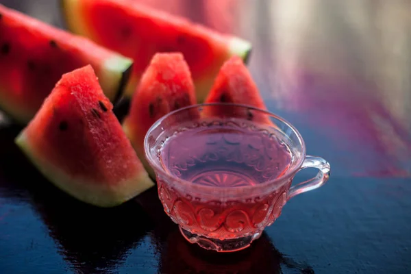 Mouth-watering watermelon tea in a transparent glass cup on wooden surface with watermelon pieces in triangle shape.