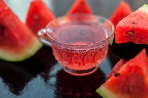 Fruit ice tea of watermelon seeds in a transparent glass cup on wooden surface along with triangular pieces of watermelon.