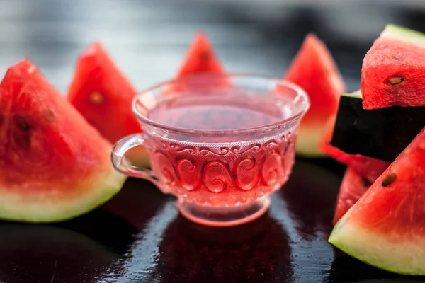 Fruit ice tea of watermelon seeds in a transparent glass cup on wooden surface along with triangular pieces of watermelon.