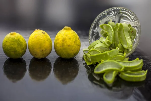 Raw cut aloevera or aloe vera gel in a glass bowl and some fresh ripe lemons or limbu or nimbo on wooden surface along with their reflection on the surface.