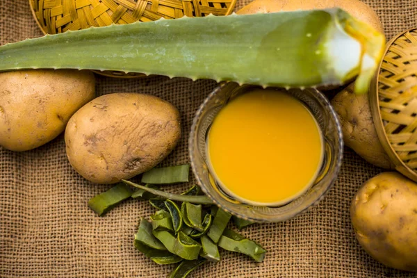 potatoes juice well mixed with aloe vera gel in glass bowl along raw potatoes and aloe vera leaves on jute bag surface, concept  of homemade conditioner and shampoo for dandruff and scalp cleaning