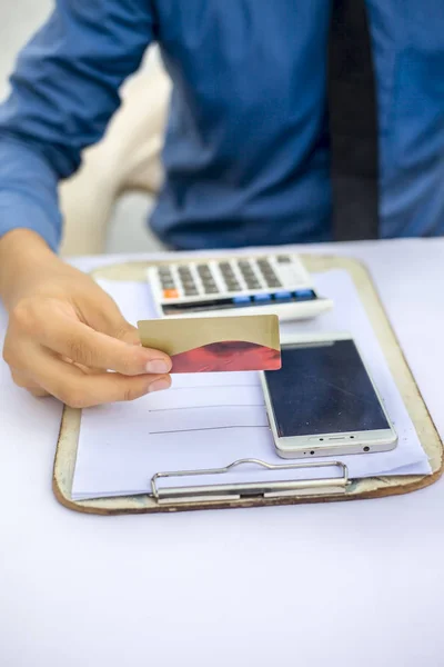 Close up of employee wearing shirt and necktie holding credit card in hand while sitting at desk with smartphone and documents on surface. Concept of banking and shopping