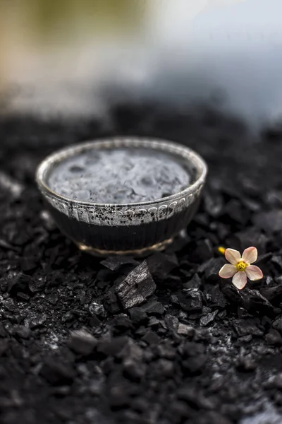 Close up of activated charcoal in a glass bowl on the wooden surface along with some raw powder of charcoal or coal spread on the surface.