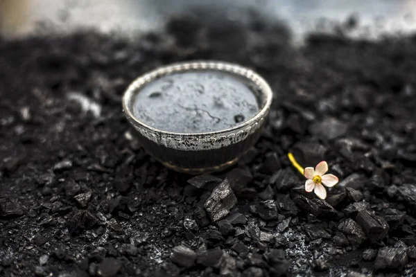 Close up of activated charcoal in a glass bowl on the wooden surface along with some raw powder of charcoal or coal spread on the surface.