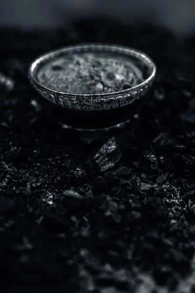 Close up of activated charcoal in a glass bowl on the wooden surface along with some raw powder of charcoal or coal spread on the surface.