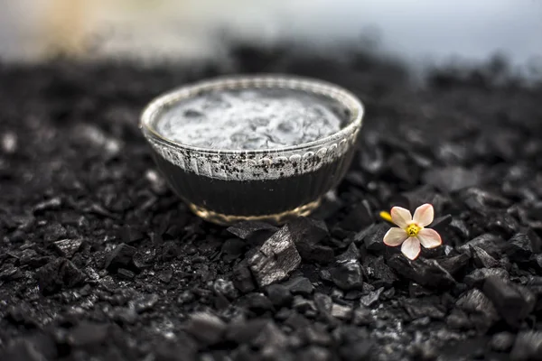 Close up of activated charcoal in a glass bowl on the wooden surface along with some raw powder of charcoal or coal spread on the surface.