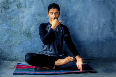 Genç çocuk siyah renkli kıyafetleri giyen ve renkli geleneksel paspas yoga yapıyor ve kaba bir mavi renkli duvara karşı Mirgi mudra veya geyik mühür pranayama yapıyor. Yatay atış.