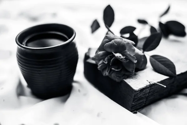 Vintage old book on white fabric with single pink colored rose and coffee in clay mug, black and white