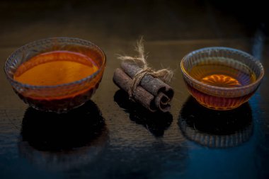 Ahşap bir yüzeyde, cam bir kasede altın ışıklı tarçın ve bal rengi saç maskesi. Yüzeyde çiğ tarçın çubuğu ve bal var..