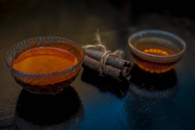 Ahşap bir yüzeyde, cam bir kasede altın ışıklı tarçın ve bal rengi saç maskesi. Yüzeyde çiğ tarçın çubuğu ve bal var..