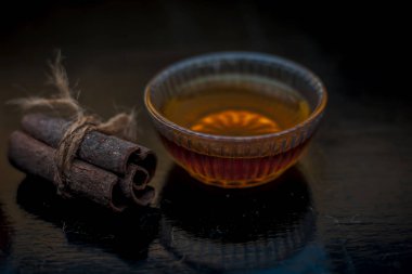 Ahşap bir yüzeyde, cam bir kasede altın ışıklı tarçın ve bal rengi saç maskesi. Yüzeyde çiğ tarçın çubuğu ve bal var..