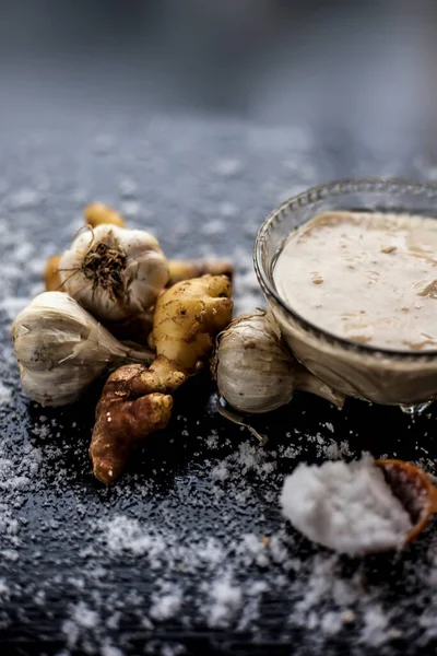 Shot of freshly ground garlic and ginger paste in a glass bowl on a black surface along with raw ginger, and some garlic with salt on sprinkle on the surface. Lasan adrak ka paste in a glass bowl on wooden surface.