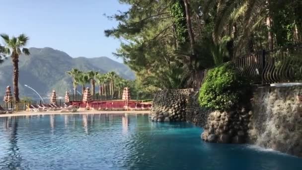 游泳池 有海水色的水 低矮的墙壁 水从里面流出来 棕榈树 远处的阳光 大海和高山 — 图库视频影像
