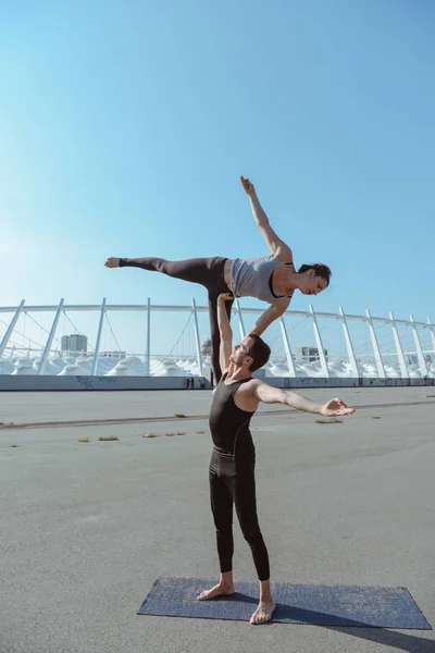 Young athletic couple practicing acroyoga at stadium urban style.Balancing in pair. Fit active pair yoga time.Sporty handsome man supporting slim beautiful brunette woman.Training time.Asana