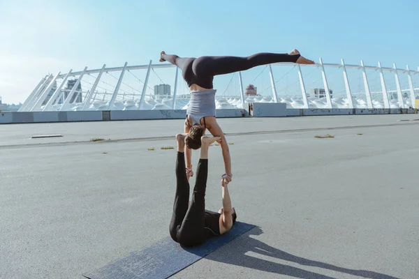 Young athletic couple practicing acroyoga at stadium urban style.Balancing in pair. Fit active pair yoga time.Sporty handsome man supporting slim beautiful brunette woman.Training time.Asana