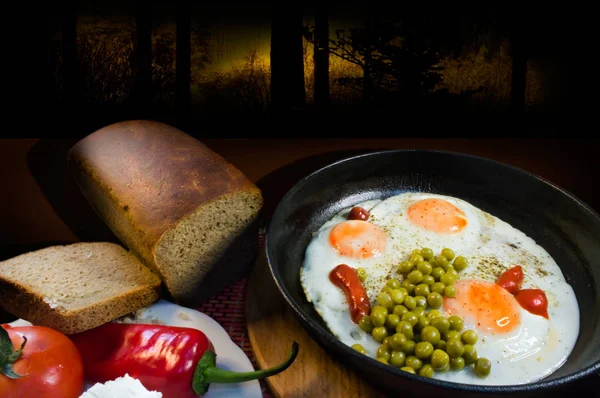 Provincial breakfast with fried eggs in a pan, peas and ketchup, as well as tomatoes, cheese, bread and pepper on a dark background.